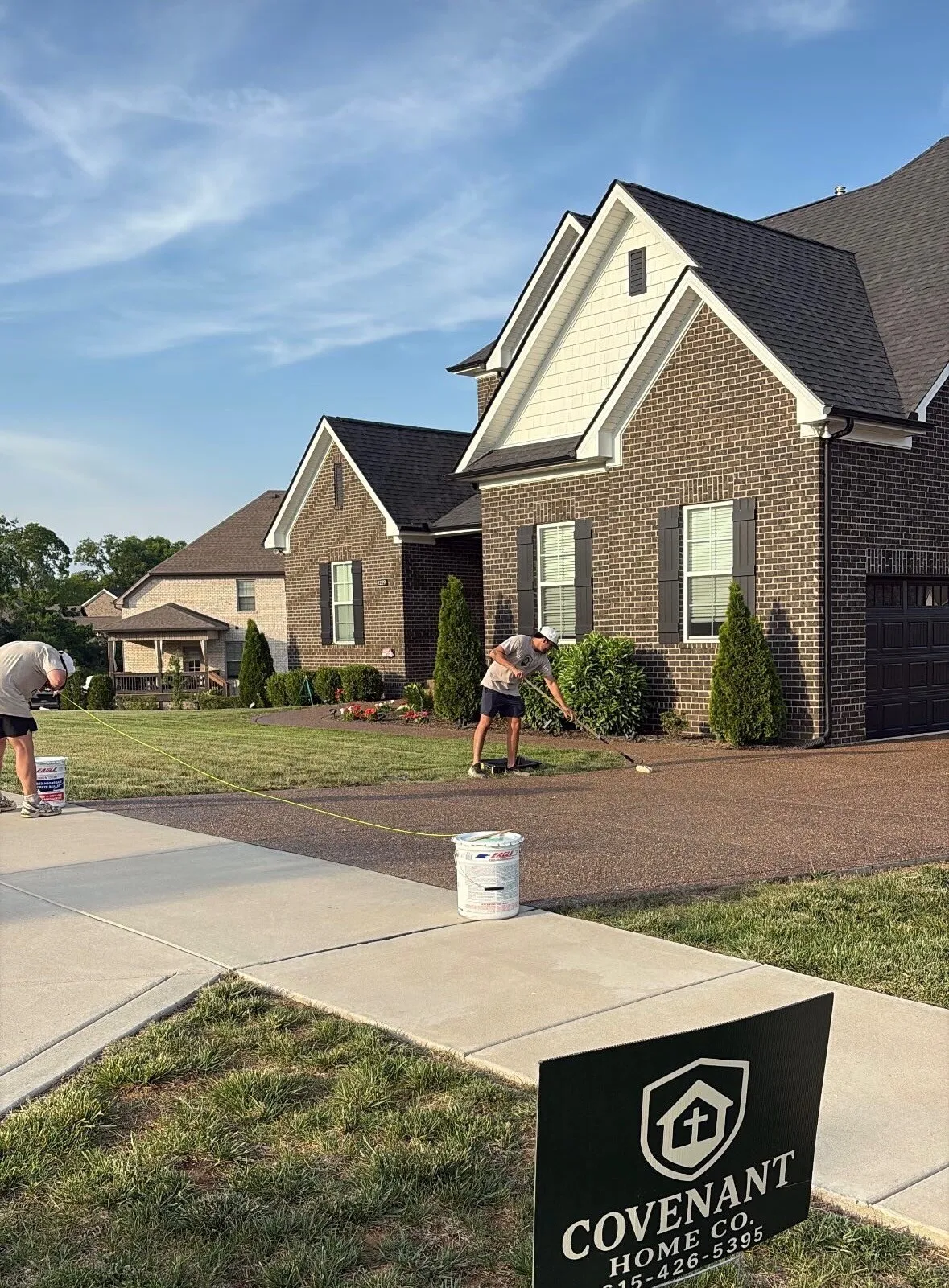 Two Covenant crew members sealing a driveway in front of a brick home, with a Covenant yard sign in the lawn